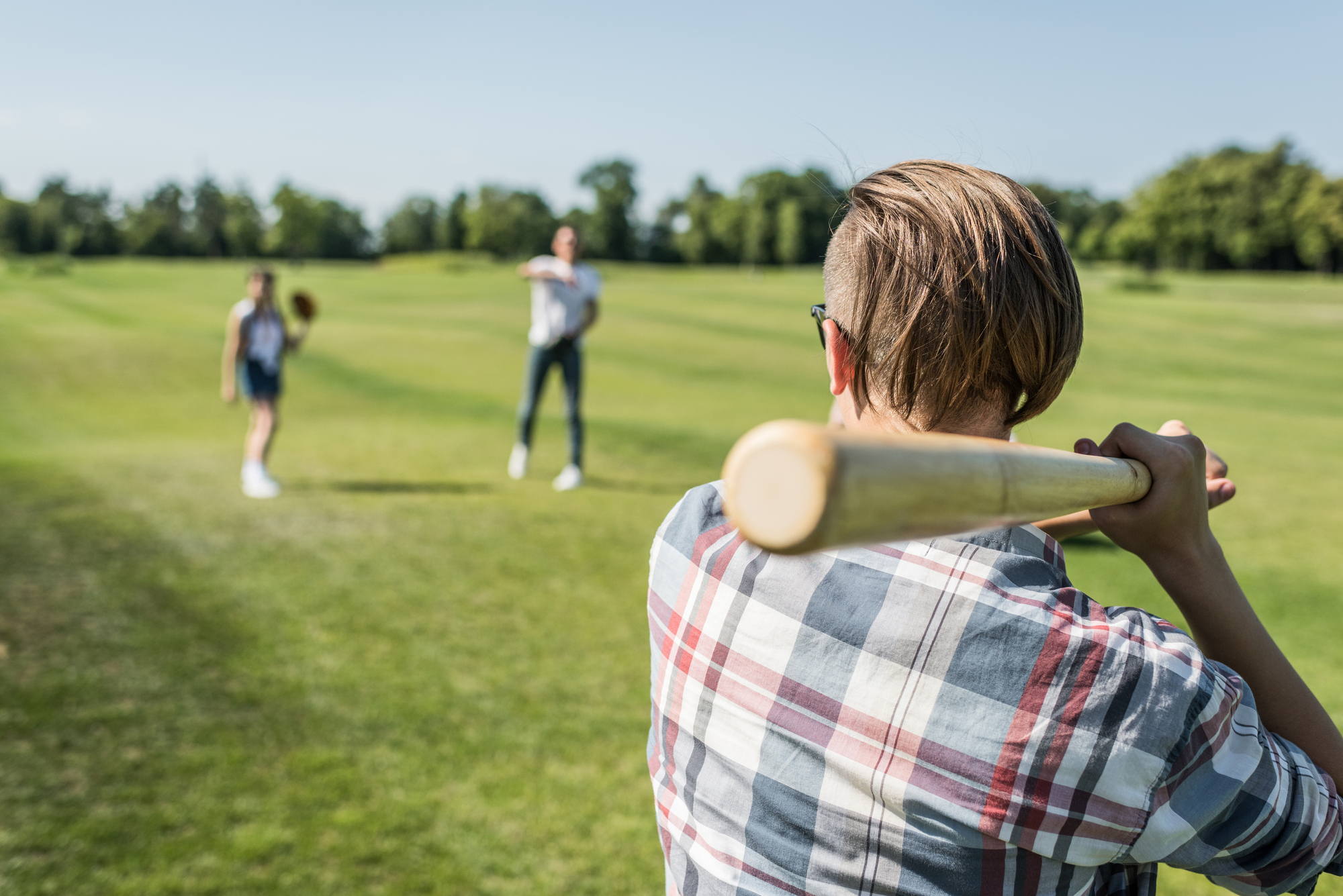 A person in a plaid shirt holds a baseball bat, preparing to hit a ball in a grassy field. Another person stands farther away, ready to catch, while a third individual watches from a distance. Trees are visible in the background under a clear sky.