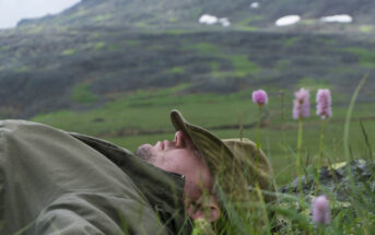 A person wearing a green jacket and hat lies on the grass in a mountainous area. The background features a blurred view of green hills with patches of snow, and pink wildflowers are in the foreground.