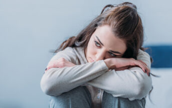 A woman with long dark hair sits with her arms resting on her knees, looking down with a pensive expression. She is wearing a light gray sweater and blue jeans. The background is blurred, giving a sense of contemplation or concern.