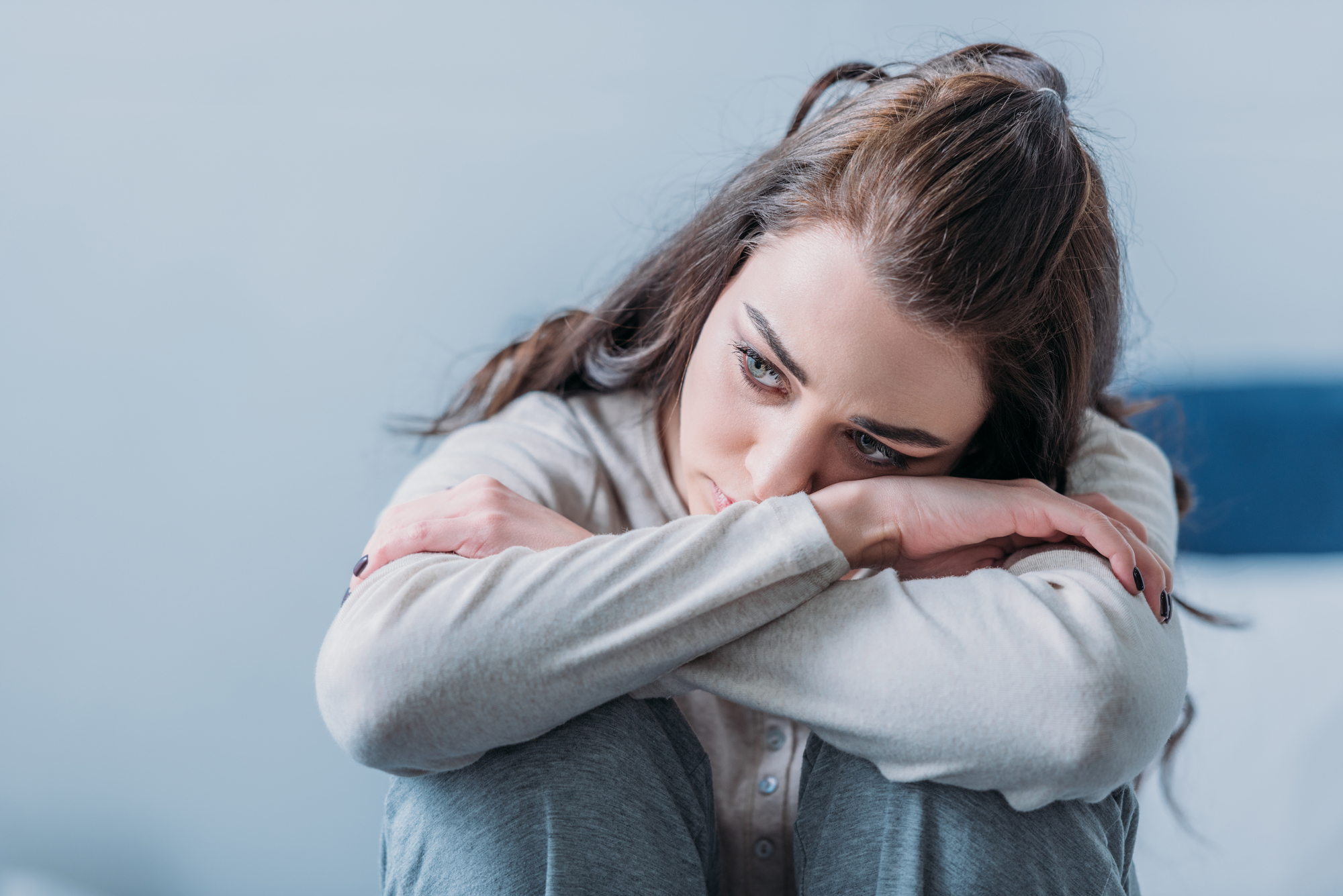 A woman with long dark hair sits with her arms resting on her knees, looking down with a pensive expression. She is wearing a light gray sweater and blue jeans. The background is blurred, giving a sense of contemplation or concern.