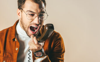 A man with glasses and a beard is angrily yelling into a phone receiver. He is wearing a white turtleneck and a brown jacket. The background is plain and neutral.