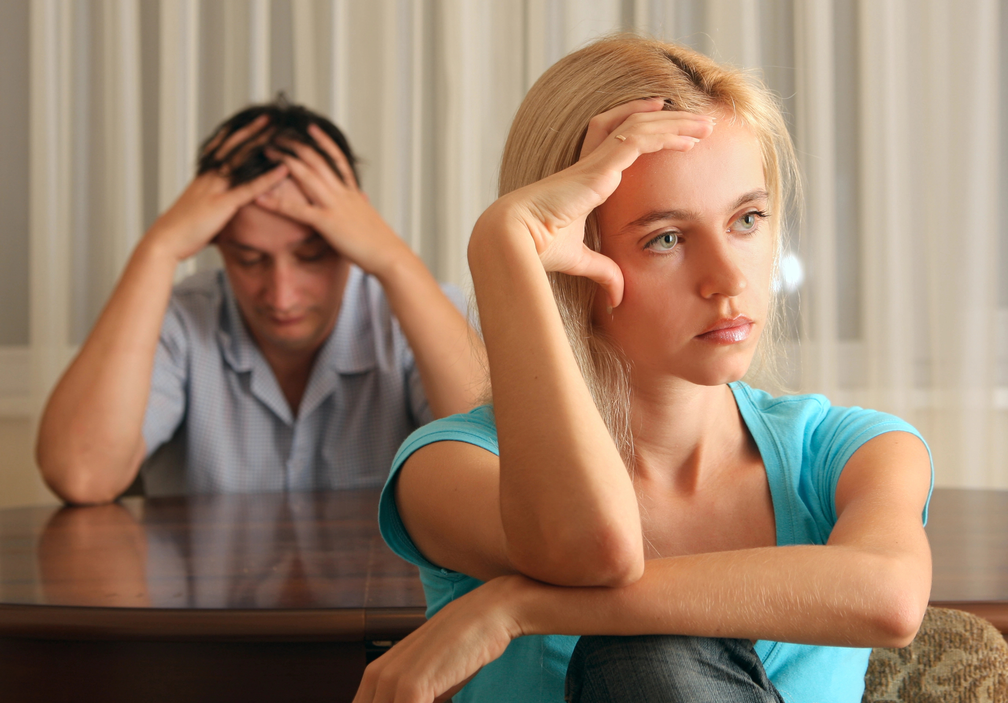 A woman with blonde hair and a blue shirt sits in the foreground looking thoughtful and slightly distressed, while a man in the background sits at a table with his head in his hands, appearing frustrated. Both are indoors with sheer curtains in the background.