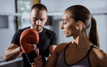 A woman with a ponytail, dressed in a tank top, holds up a red boxing glove while a man with a trimmed beard and short hair, wearing a black shirt, adjusts it in a gym setting. The background shows a blurred view of gym equipment and a large window.