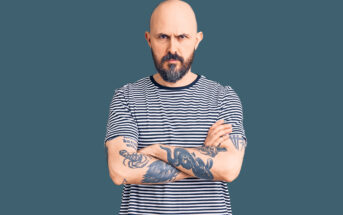 A bald man with a beard and tattoos on both arms stands with his arms crossed, wearing a black and white striped shirt. The background is a solid dark teal color. He has a serious expression on his face.