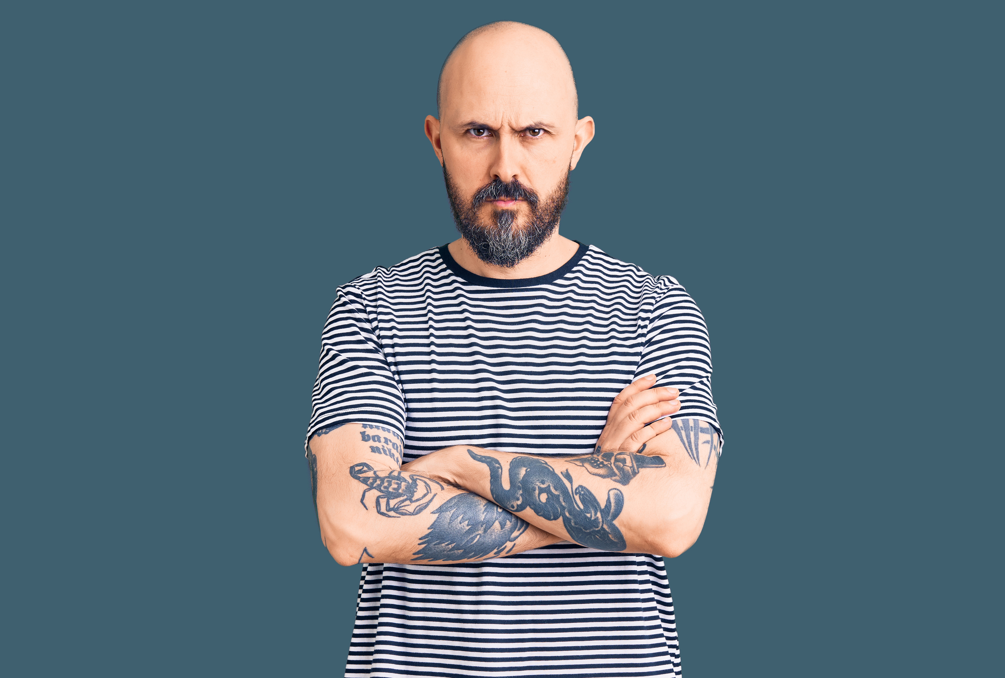 A bald man with a beard and tattoos on both arms stands with his arms crossed, wearing a black and white striped shirt. The background is a solid dark teal color. He has a serious expression on his face.