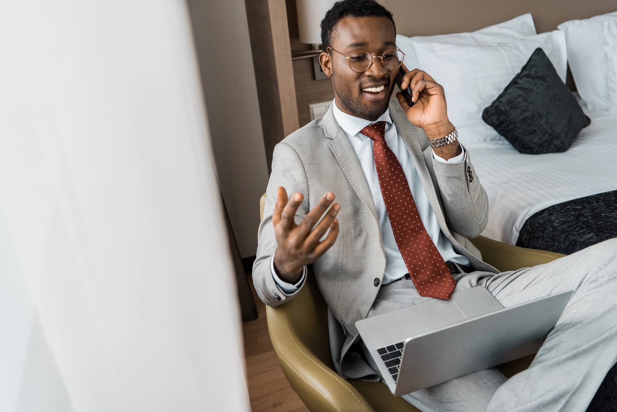 A man in a gray suit and red tie sits on a chair with a laptop on his lap, smiling and talking on the phone. He appears to be in a modern, well-lit room with a neatly made bed in the background.