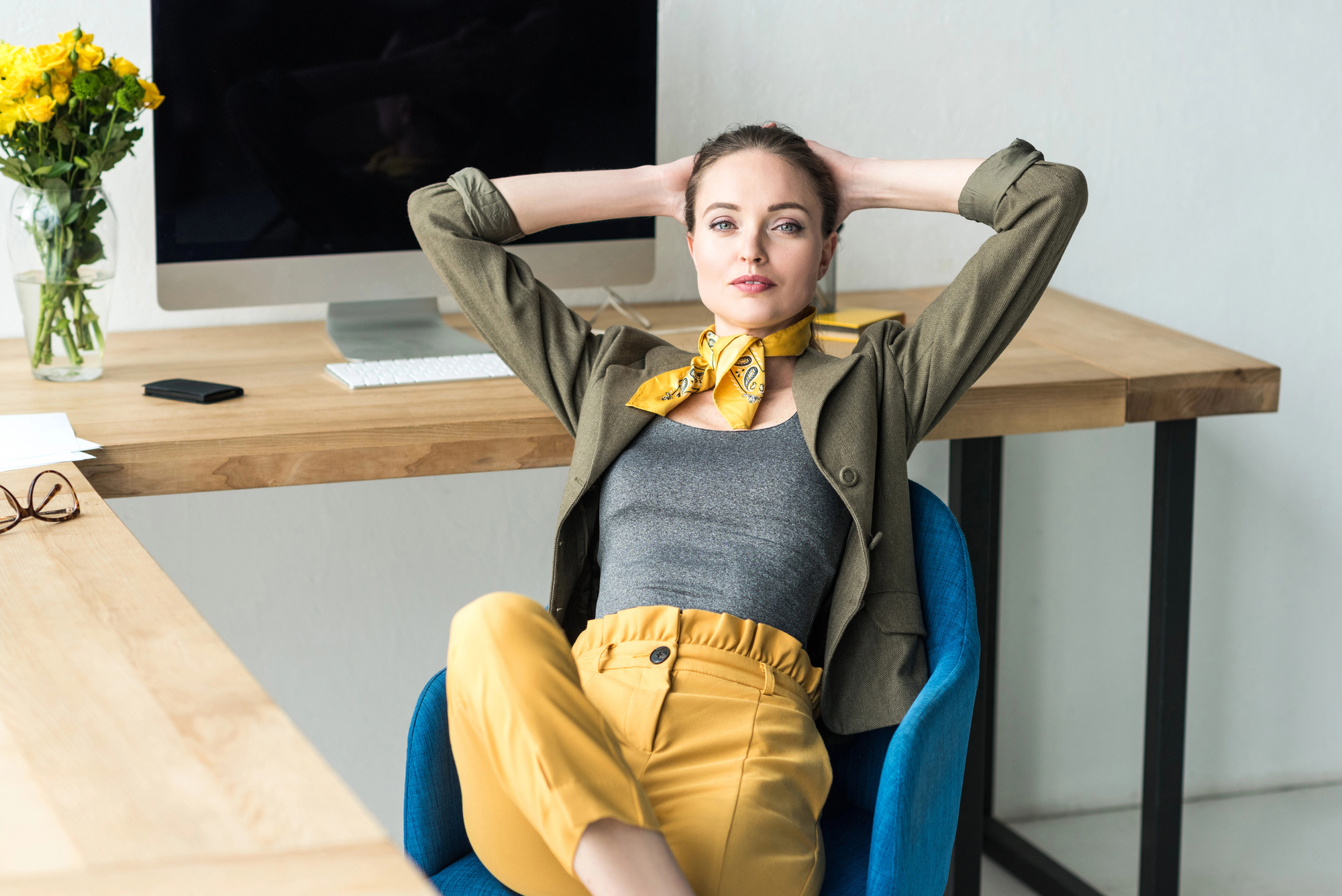 A woman sits relaxed in an office chair, hands behind her head, in front of a desk with a computer, keyboard, and flowers. She wears a blazer, gray top, yellow pants, and a matching scarf. The scene has a modern, minimalist vibe.