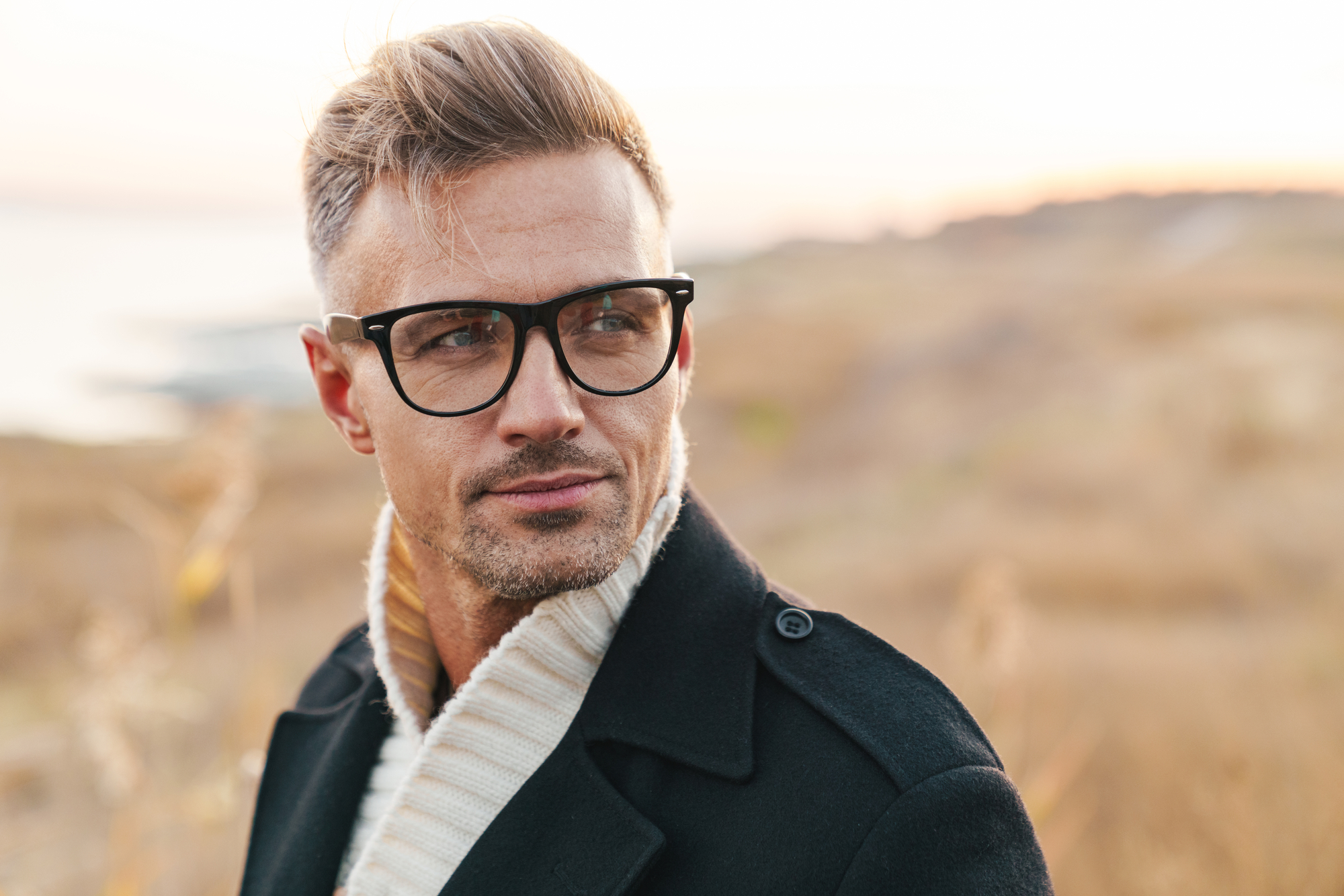 A man with blond hair and glasses is wearing a black coat and a cream-colored scarf. He is outdoors in a natural setting with dry grass and blurred landscape in the background. The lighting suggests that it is either early morning or late afternoon.