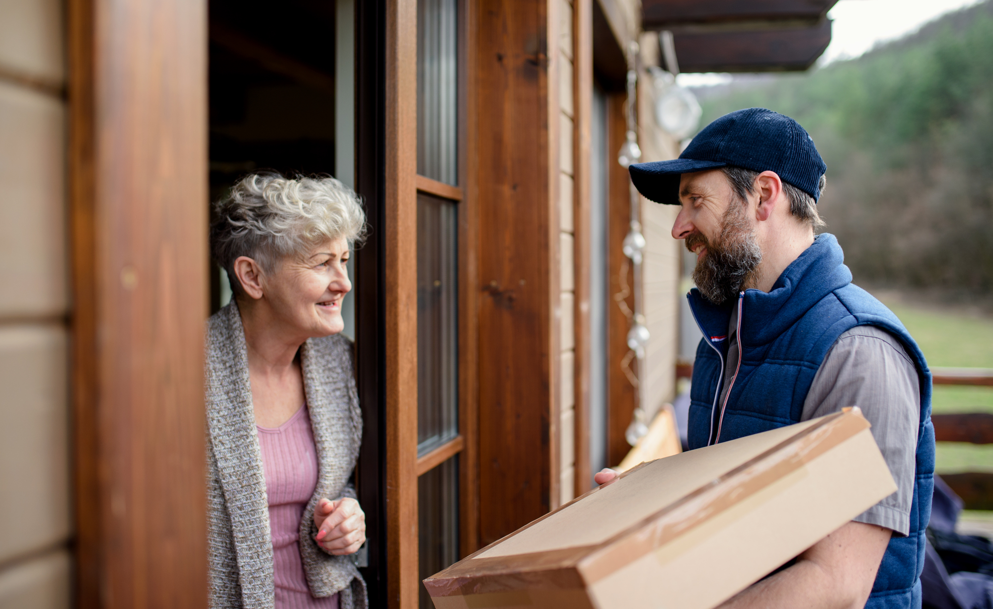 An older woman with short gray hair is receiving a package from a delivery person at her doorstep. The delivery person, wearing a blue cap and vest, is holding a large cardboard box and smiling at the woman. The background shows a wooden house and a garden.