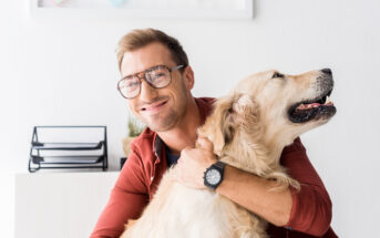 A smiling man with glasses hugs a happy Golden Retriever in a cozy indoor setting. The background shows a white wall with framed pictures and a desk organizer. The man wears a red jacket and a watch, while the dog looks content and relaxed.