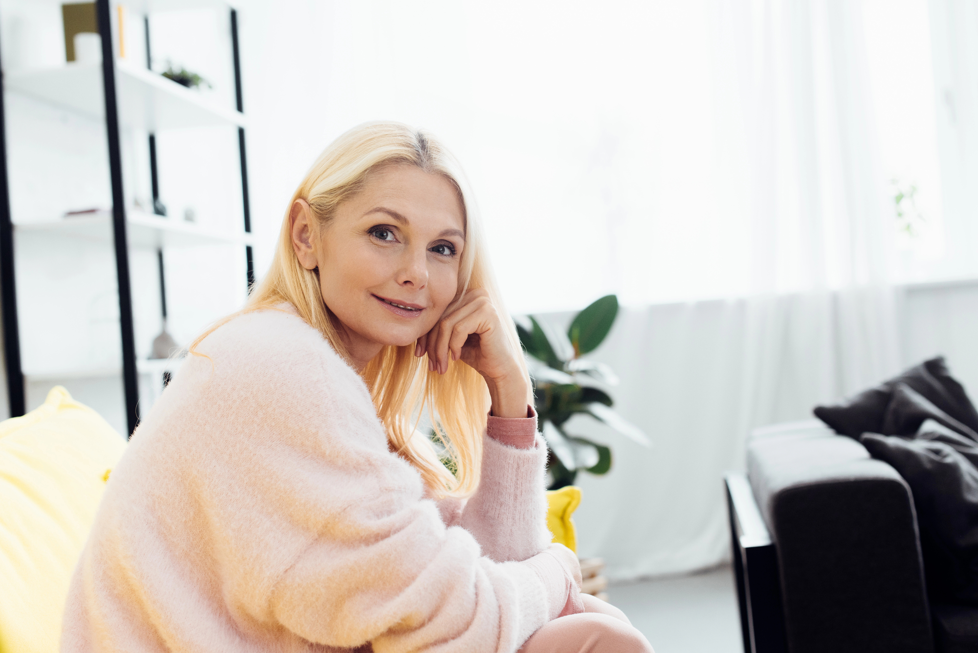 A blonde woman in a light pink sweater is sitting on a yellow chair, resting her head on her hand and smiling. She is in a well-lit room with white walls, a shelf with books and plants in the background, and a grey sofa with a cushion.