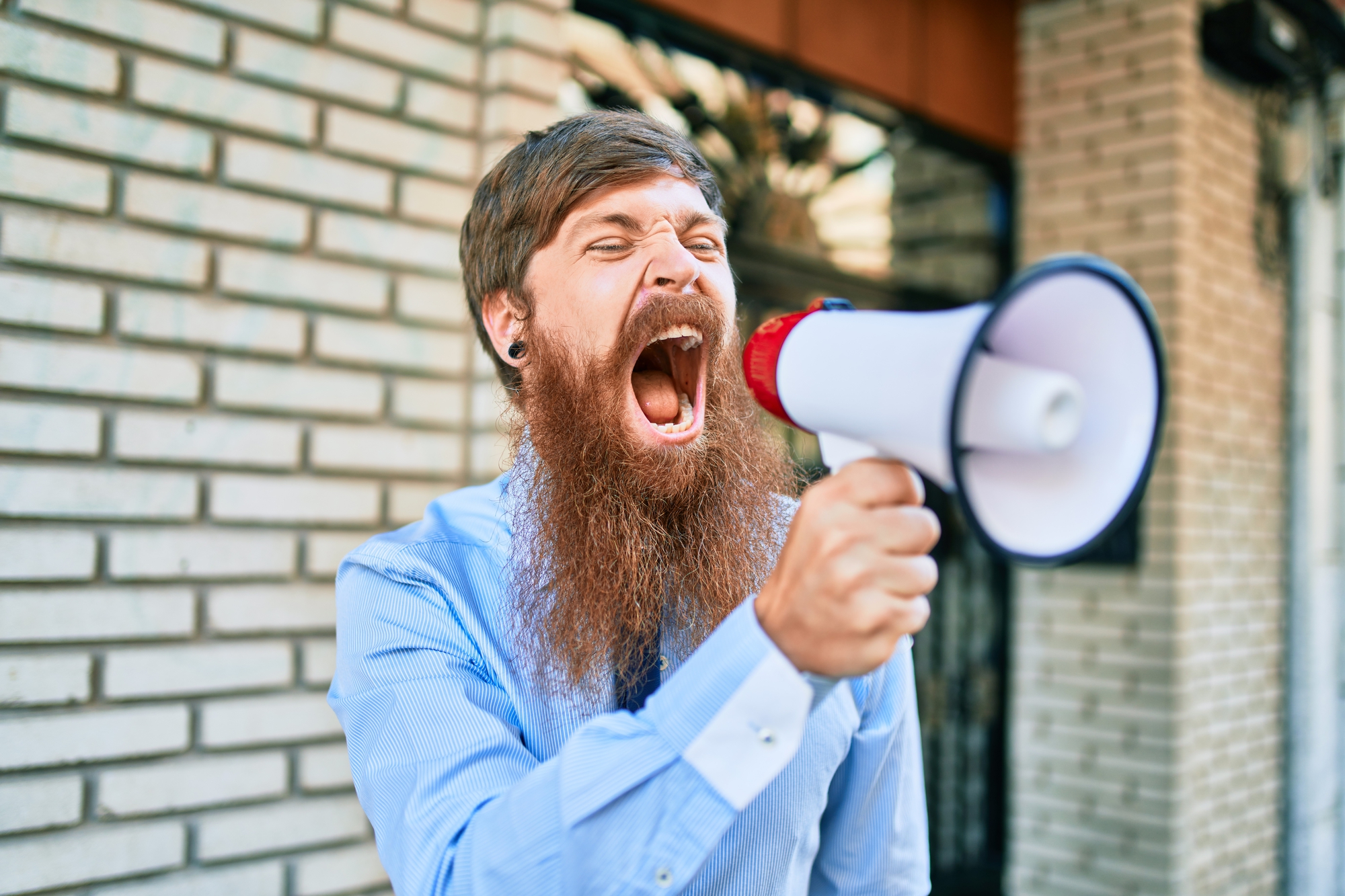 A bearded man in a blue shirt shouts into a megaphone while standing against a brick wall. His expression is intense and determined, and he appears to be passionately making an announcement or protest.