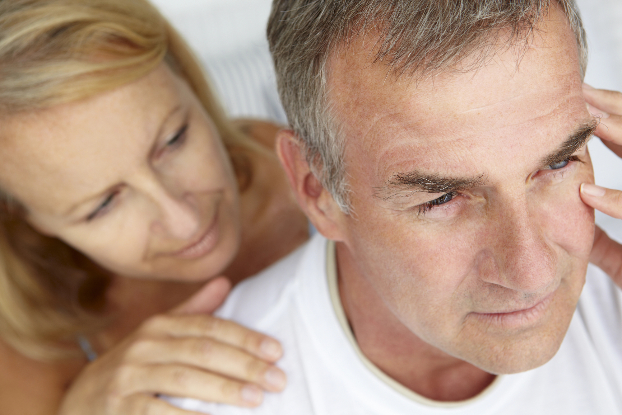 A close-up of an older couple. The man is in the foreground, looking contemplative with his hand near his temple. The woman is slightly behind him, with her hand gently resting on his shoulder, looking at him with concern. Both are wearing casual clothing.