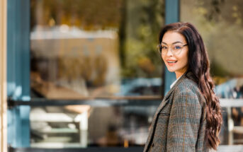 A woman with long, dark hair and glasses, dressed in a plaid coat, smiles while standing outside near a blue-framed glass window. The background shows a blurred view of trees and buildings. She is turning slightly towards the camera.