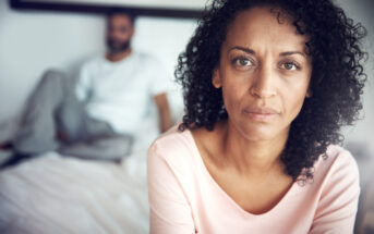 A woman with curly hair and a serious expression is sitting on the edge of a bed, wearing a pink top. In the background, a man with a beard and casual clothing is sitting on the bed, slightly blurred. The setting appears to be a bedroom.