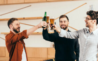 Three men cheerfully clink glasses in a modern bar. Two hold beer mugs, and one holds a beer bottle. They are smiling and appear to be enjoying their time together. The background features light wooden decor with minimalist designs.