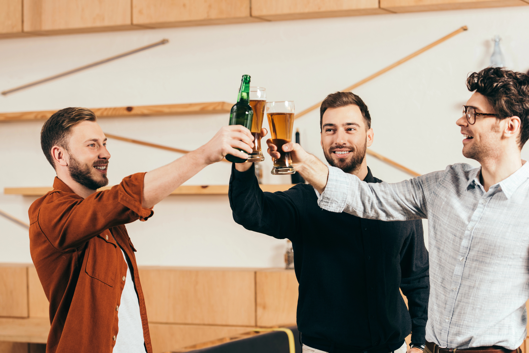 Three men cheerfully clink glasses in a modern bar. Two hold beer mugs, and one holds a beer bottle. They are smiling and appear to be enjoying their time together. The background features light wooden decor with minimalist designs.