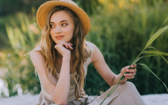 A young woman with long, wavy hair sits outdoors on a sunny day, wearing a straw hat and a beige outfit. She gazes thoughtfully to the side, resting her chin on her hand, and holds a long blade of grass in her other hand. Green foliage is visible in the background.