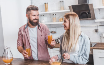 A bearded man and a blonde woman smile at each other while holding glasses of orange juice in a modern kitchen. The woman is also holding a mixing bowl with a whisk inside. A glass pitcher of juice is on the counter next to them.