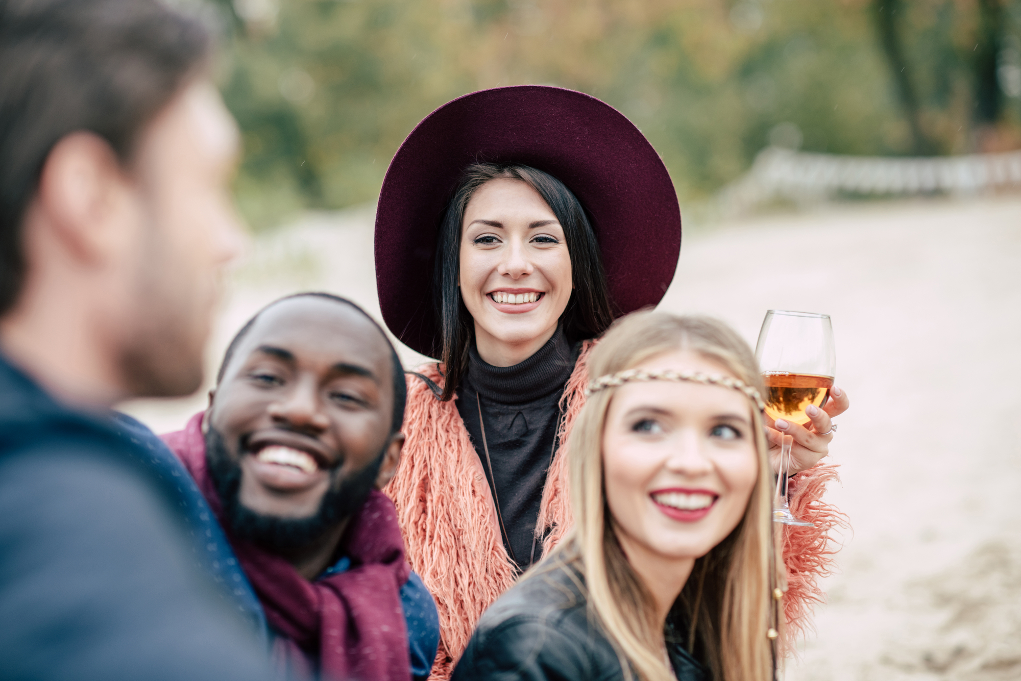 Group of friends enjoying outdoor gathering. Smiling woman in center wears dark hat and holds wine glass. Man on left and woman on right look towards man in foreground. All are cheerful, with blurred outdoor background suggesting a relaxed and casual setting.