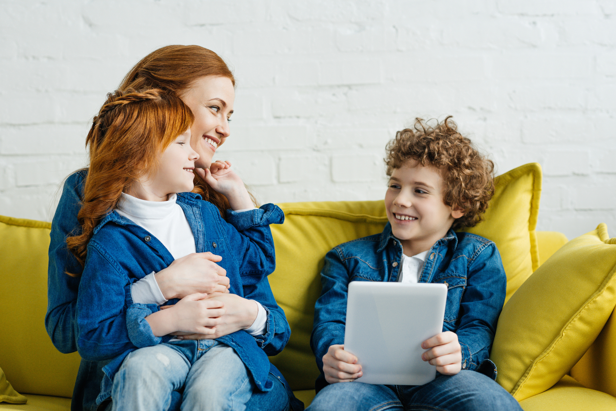 A woman with red hair embraces a red-haired child while sitting on a yellow couch. Another child with curly hair sits beside them, holding a tablet. They all wear blue denim outfits and smile at each other, creating a warm, happy scene.