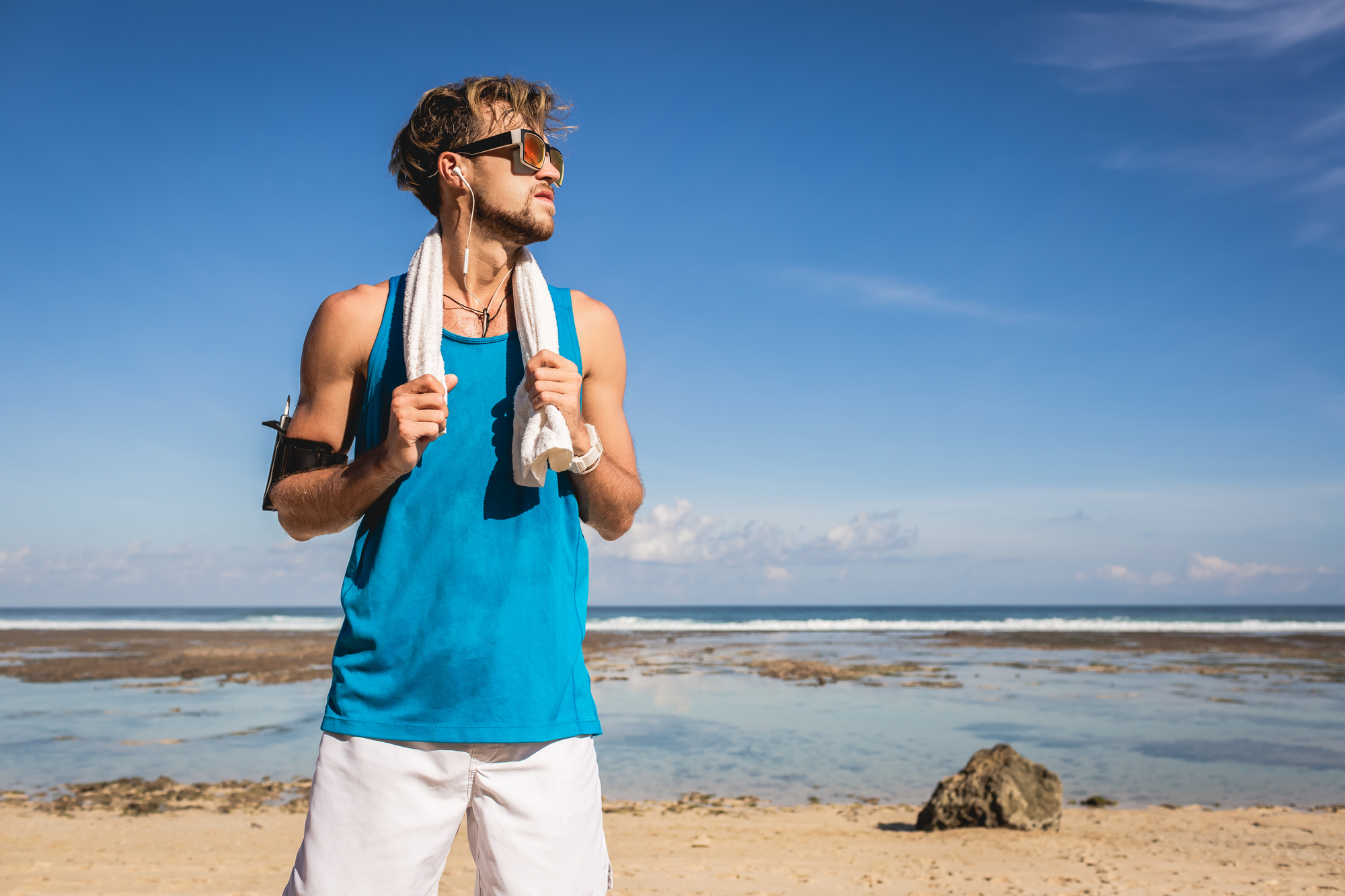 A young man wearing a blue tank top and white shorts stands on a beach. He has sunglasses on, with a white towel draped over his shoulders and earphones in his ears. The sky is clear and the ocean is visible in the background.