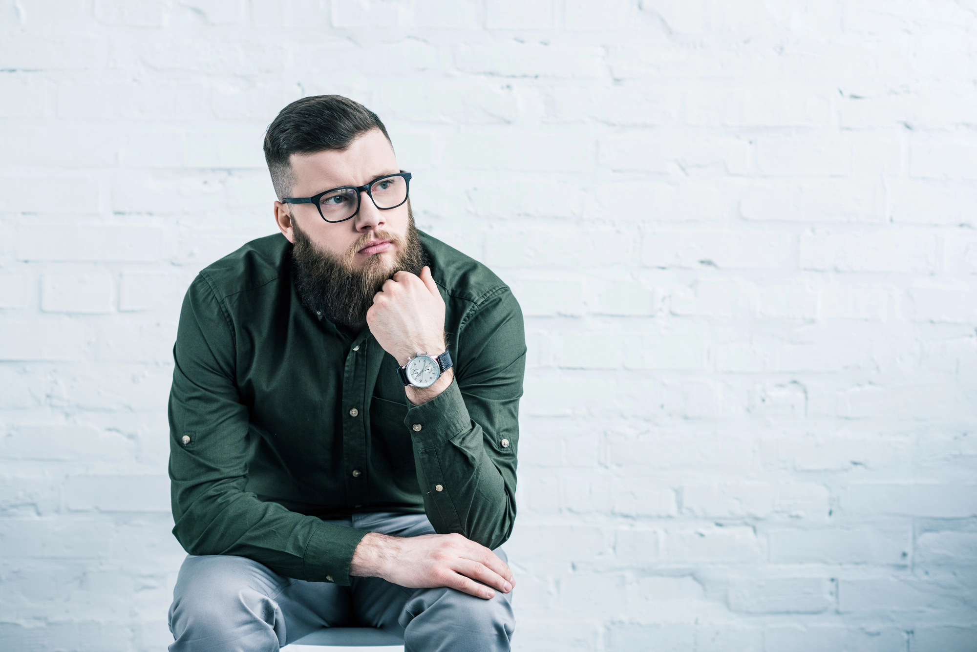 A bearded person with glasses, wearing a dark green shirt and light-colored pants, is sitting against a white brick wall. They are resting their chin on one hand and looking thoughtfully into the distance. A wristwatch is visible on their left wrist.