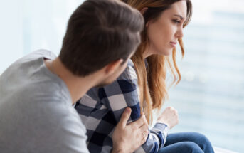 A man with short brown hair and a grey shirt sits beside a woman with long wavy brown hair and a checkered shirt. The man is gently touching the woman's arm as she looks away, appearing thoughtful or upset. They are indoors with a blurred background.