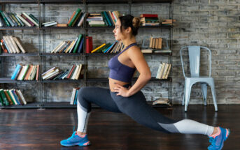 A woman in athletic wear performs a lunge exercise in front of a bookshelf with scattered books. She wears a blue sports bra, gray leggings, and blue athletic shoes. A metal chair is positioned against the brick wall background.