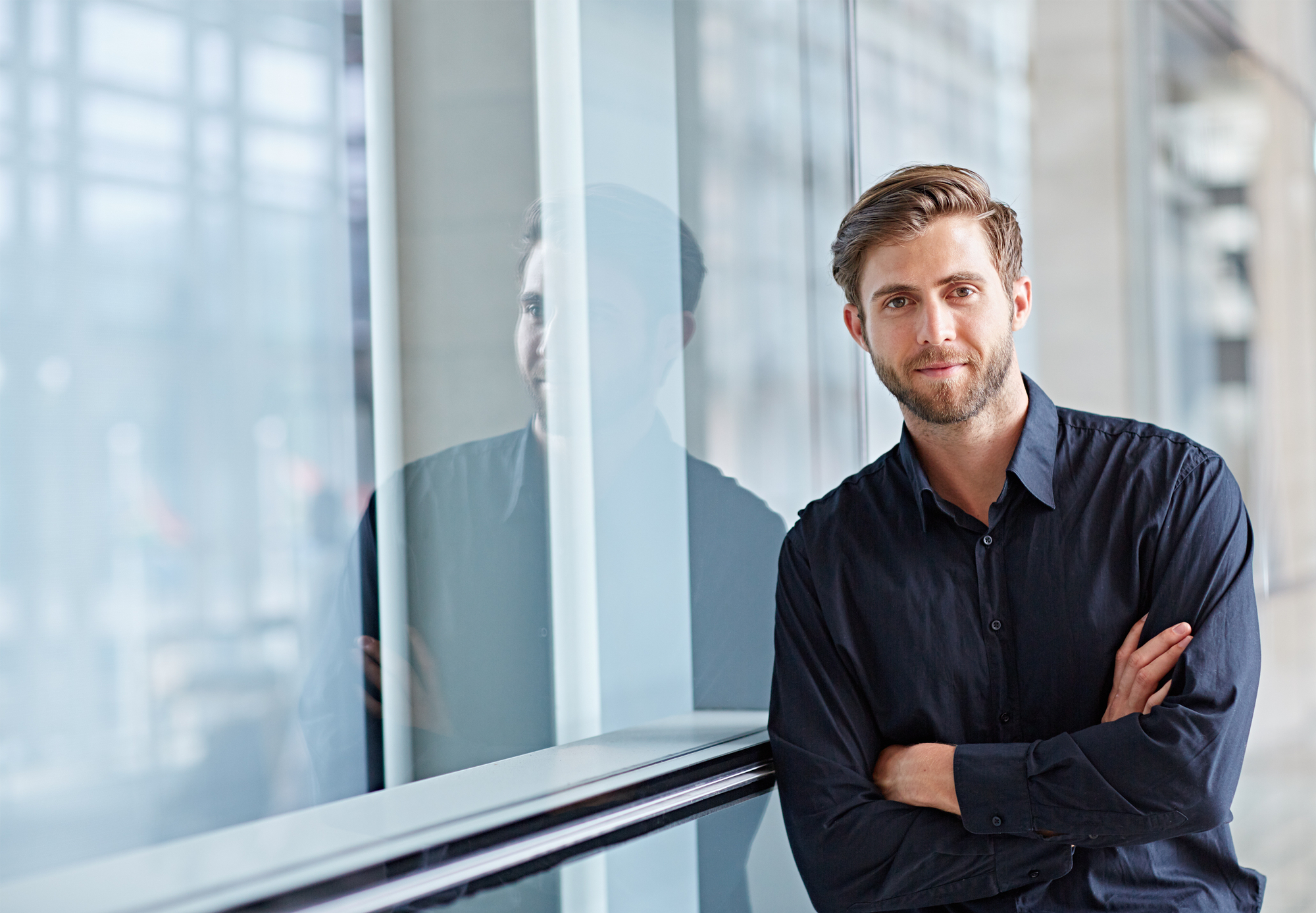 A man with a beard, wearing a dark shirt, stands with his arms crossed next to a large window. He is looking at the camera with a slight smile. The window creates a faint reflection of him, and the background appears to be a modern indoor setting.