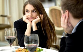 A woman with long brown hair, resting her face in her hands, looks thoughtfully to the side while sitting at a table with a man. They are in a restaurant, and there are wine glasses and plates of food in front of them.