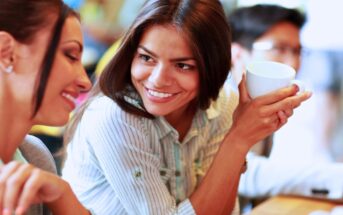 Two women are seated at a table, engaged in a friendly conversation. One woman is holding a white cup and smiling at the other, who is also smiling. The setting is lively, and there are blurred figures and soft-focus greenery in the background.