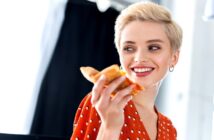 young woman with short blonde hair smiling while holding a slice of pizza