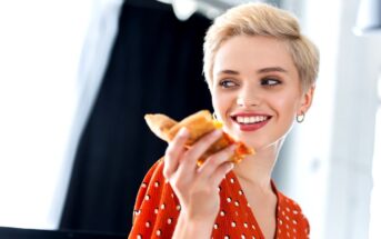 A person with short blond hair smiles while holding a slice of pizza. They are wearing a red shirt with white polka dots and earrings. The background is blurred, showing a dark curtain and some light coming through a window.