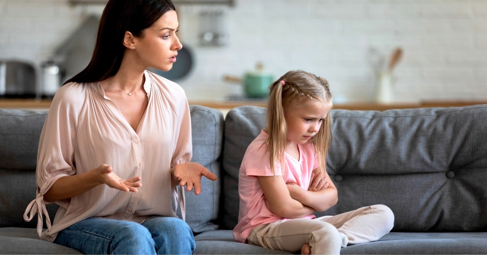 A woman with dark hair is sitting on a grey couch, speaking emotionally with her hands outstretched. Next to her, a young girl with blonde hair in braids sits with arms crossed and a pout, looking away. Both appear to be in a tense conversation.