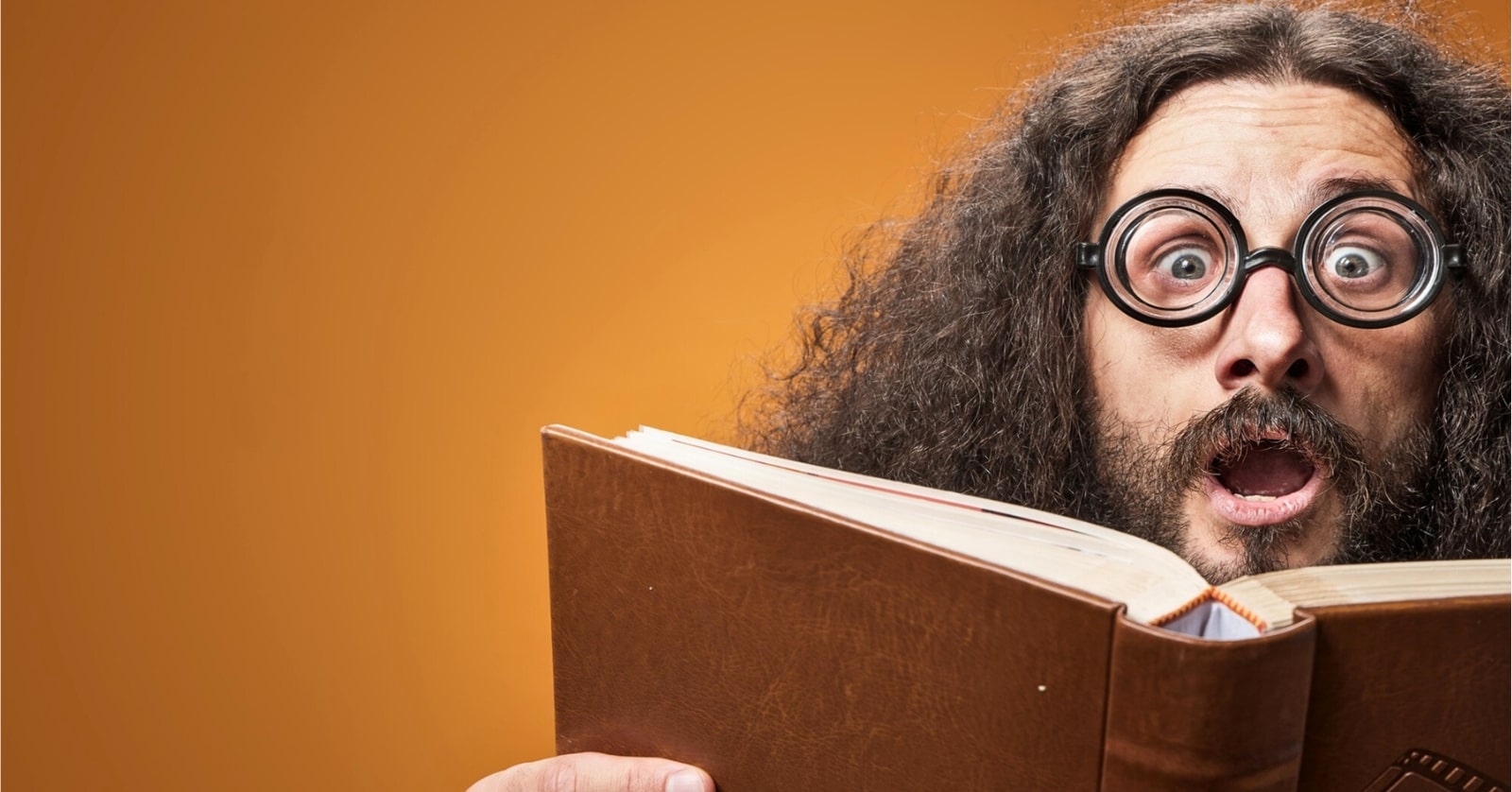 A person with long curly hair and thick-rimmed round glasses is wearing a surprised expression while holding an open book. The background is a solid orange color.