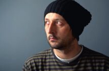 A man wearing a black knit beanie and a striped shirt looks off to the side against a gray background. He has a slight beard and appears thoughtful or contemplative.