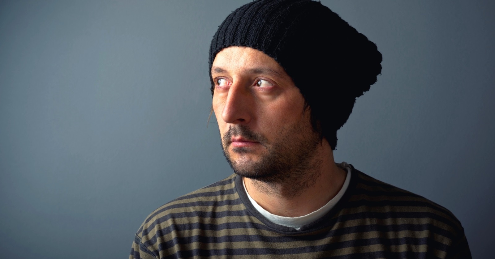 A man wearing a black knit beanie and a striped shirt looks off to the side against a gray background. He has a slight beard and appears thoughtful or contemplative.
