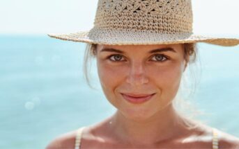 Close-up of a person smiling at the camera, wearing a wide-brimmed straw hat. The background is blurred but appears to be a calm and sunny beach with blue ocean water. The person has a relaxed and happy expression on their face.