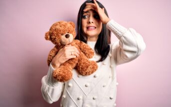 A woman with dark hair in a white sweater nervously holds a brown teddy bear. Her left hand is on her forehead, and her face shows an expression of worry or concern. She stands against a pink background.