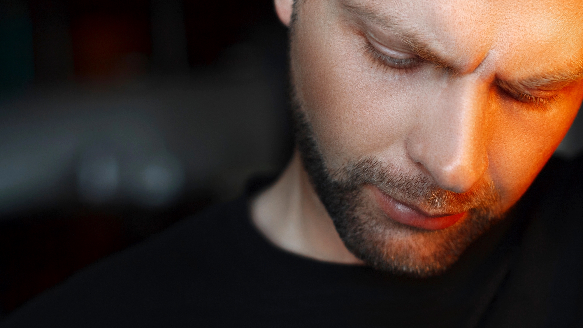 A close-up of a man with a beard and a furrowed brow looking downward. The lighting casts an orange hue on one side of his face, highlighting his contemplative or serious expression. He is wearing a black shirt and the background is blurred.