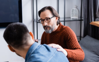 A bearded man with glasses and a red sweater sits on a couch, gently placing his hand on the shoulder of another man who is facing away, wearing a blue shirt. They appear to be having a serious conversation in a cozy, modern living room.