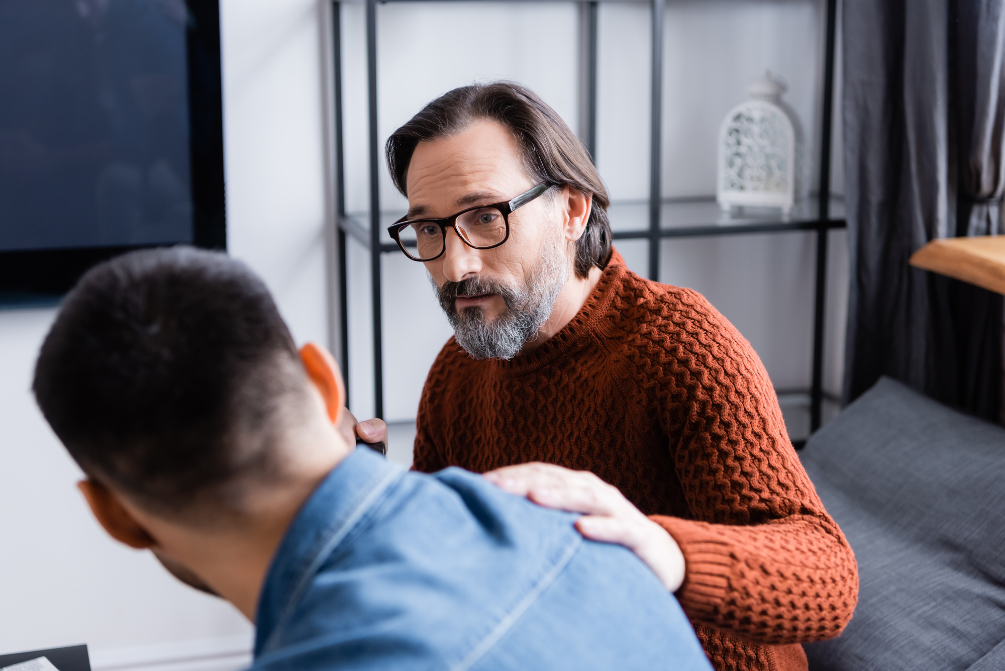 A bearded man with glasses and a red sweater sits on a couch, gently placing his hand on the shoulder of another man who is facing away, wearing a blue shirt. They appear to be having a serious conversation in a cozy, modern living room.