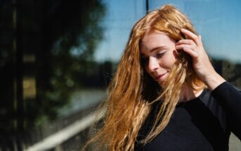 A woman with long, wavy red hair stands in front of a reflective glass surface. She is wearing a black long-sleeved top and is gently touching her hair with one hand while smiling softly. Sunlight illuminates her face and hair, creating a warm, natural glow.