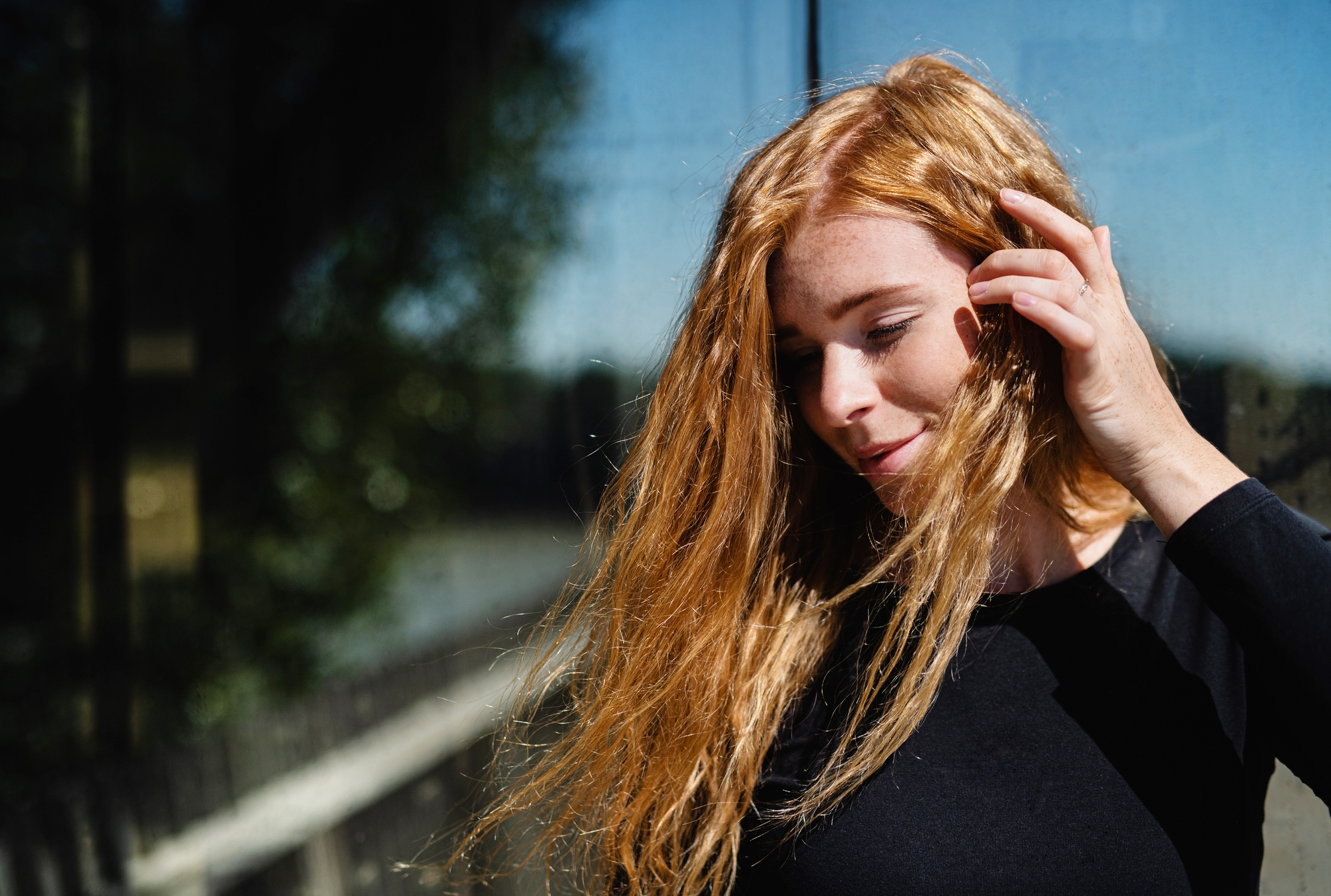 A woman with long, wavy red hair stands in front of a reflective glass surface. She is wearing a black long-sleeved top and is gently touching her hair with one hand while smiling softly. Sunlight illuminates her face and hair, creating a warm, natural glow.