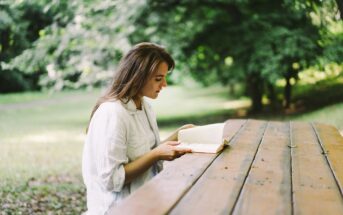 A young woman with long hair sits at a wooden picnic table outdoors, engrossed in reading a book. She is wearing a light-colored shirt, and the background features lush green trees and grass, suggesting a serene and peaceful park setting.