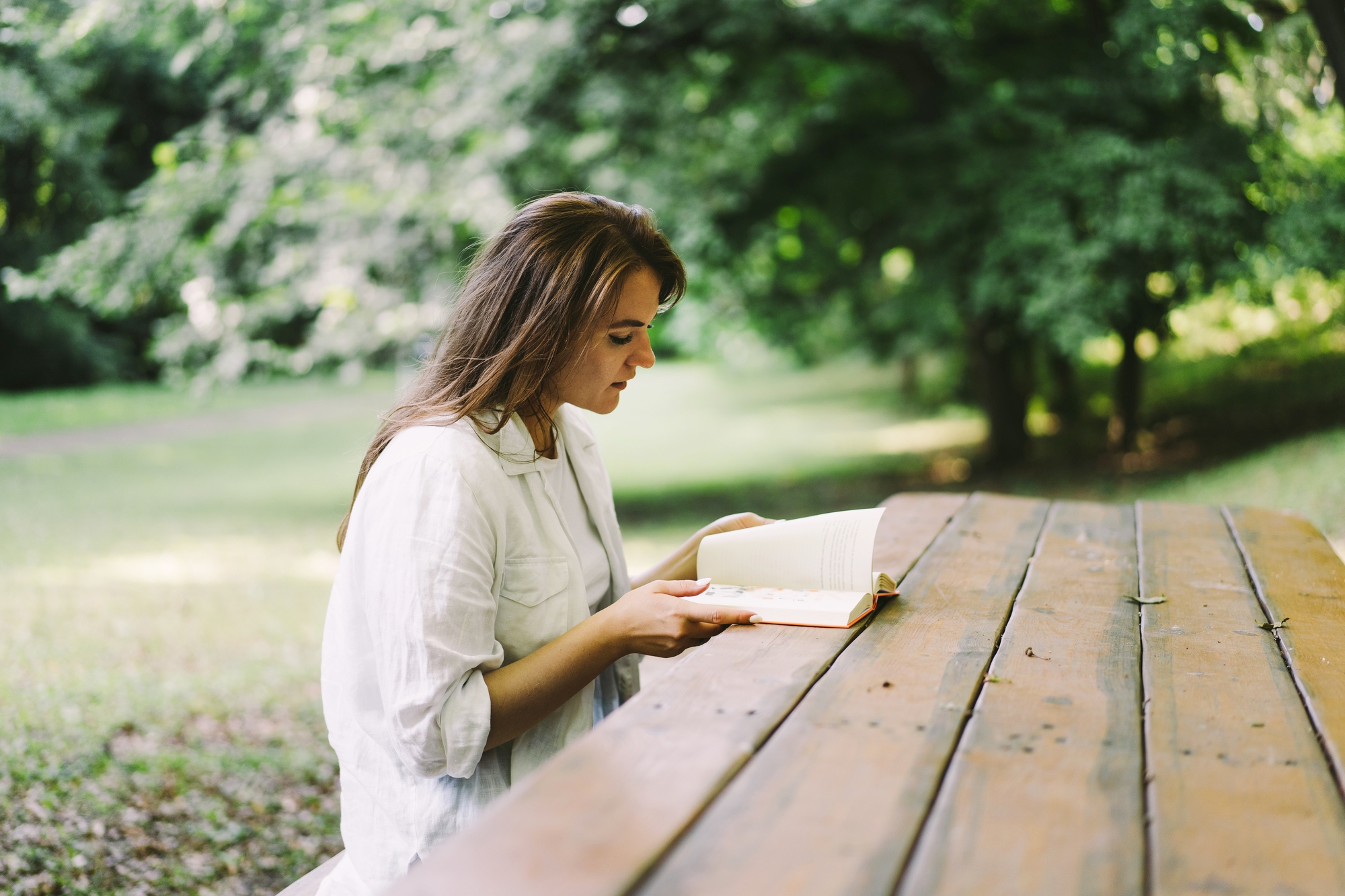 A young woman with long hair sits at a wooden picnic table outdoors, engrossed in reading a book. She is wearing a light-colored shirt, and the background features lush green trees and grass, suggesting a serene and peaceful park setting.