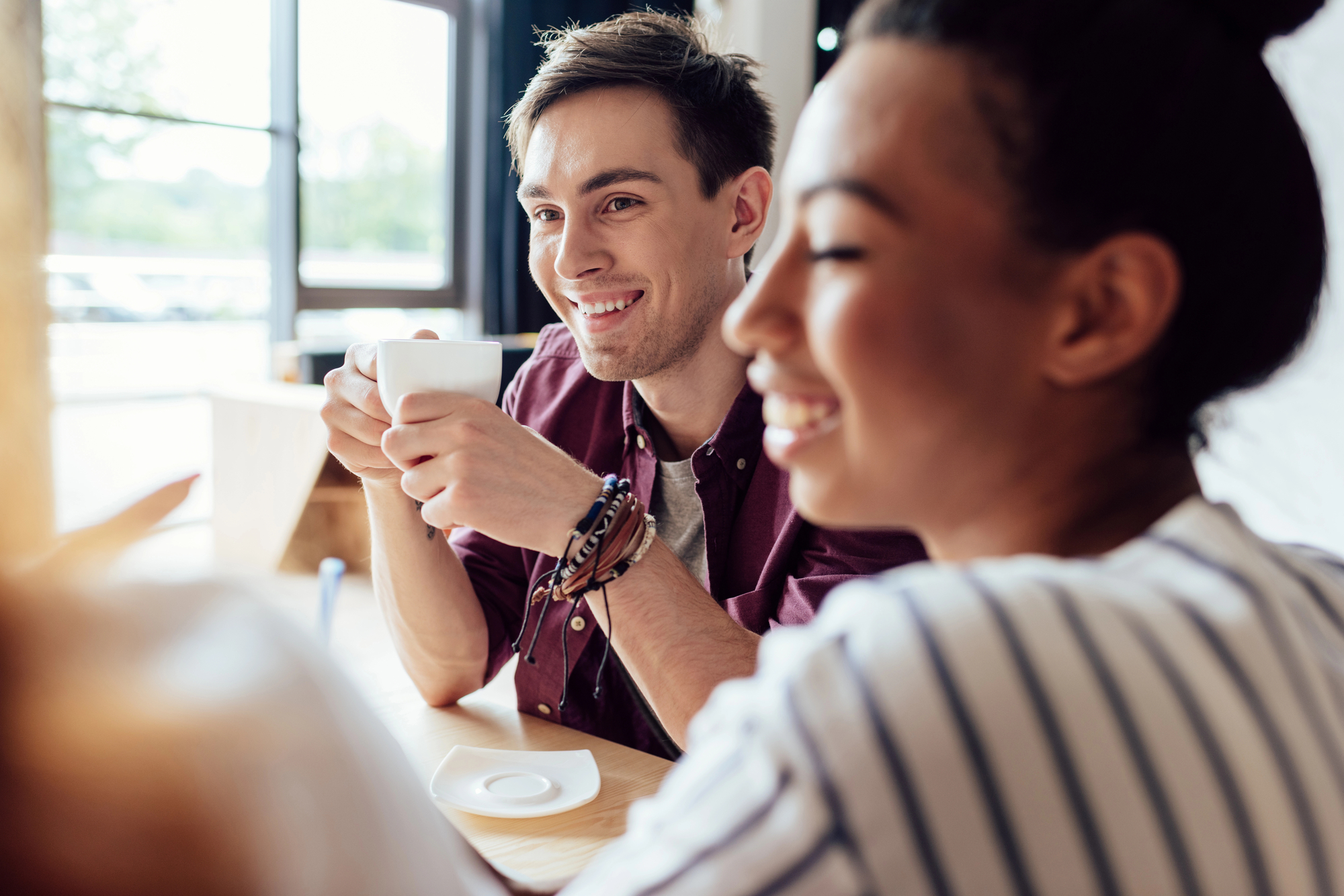 A young man and woman smile and enjoy a conversation at a bright cafe. The man holds a white coffee cup in both hands, while the woman looks at something out of the frame, smiling. They are seated at a wooden table with natural light coming in through large windows behind them.