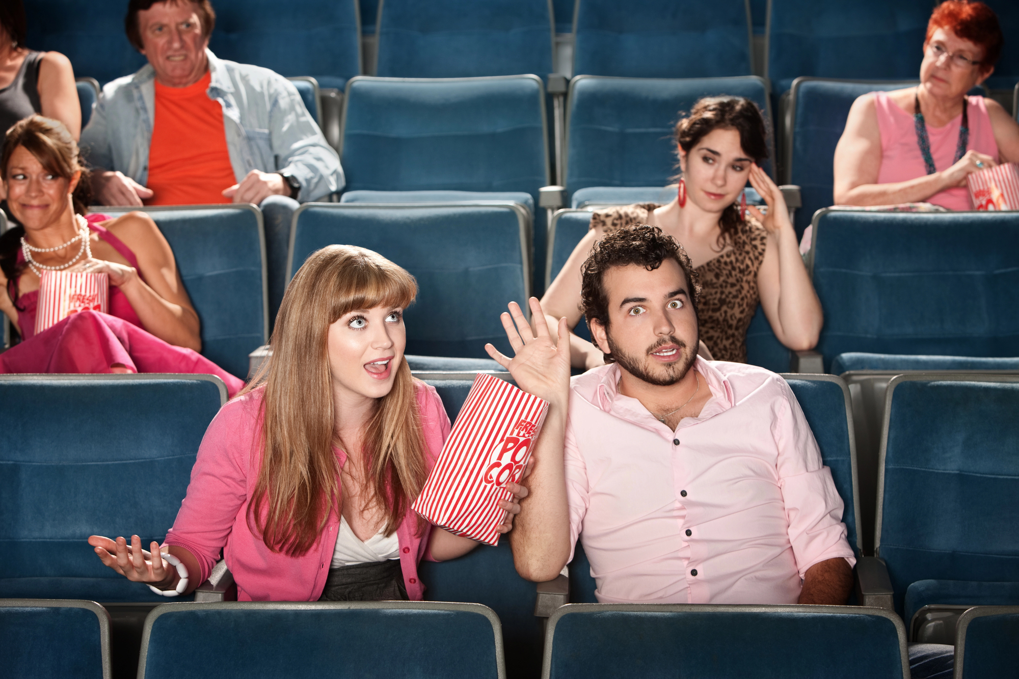 A woman and a man, both dressed in pink, sit in a movie theater with shocked expressions, holding a bag of popcorn. Other patrons in the rows behind them have varied reactions, some looking amused and others surprised. The theater seats are blue.