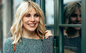 A woman with shoulder-length blonde hair smiles while standing by a window with a reflection of her visible in the glass. She is wearing a black and white patterned top and holds the edge of the window frame, exuding a cheerful and relaxed demeanor.